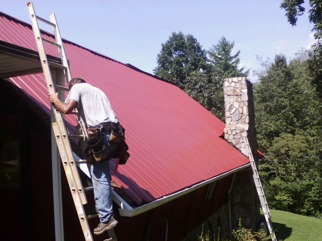 Roofer on a ladder beside a red metal roof and stone chimney — Soffit & Fascia Repair by Roofing Brothers in Panguitch